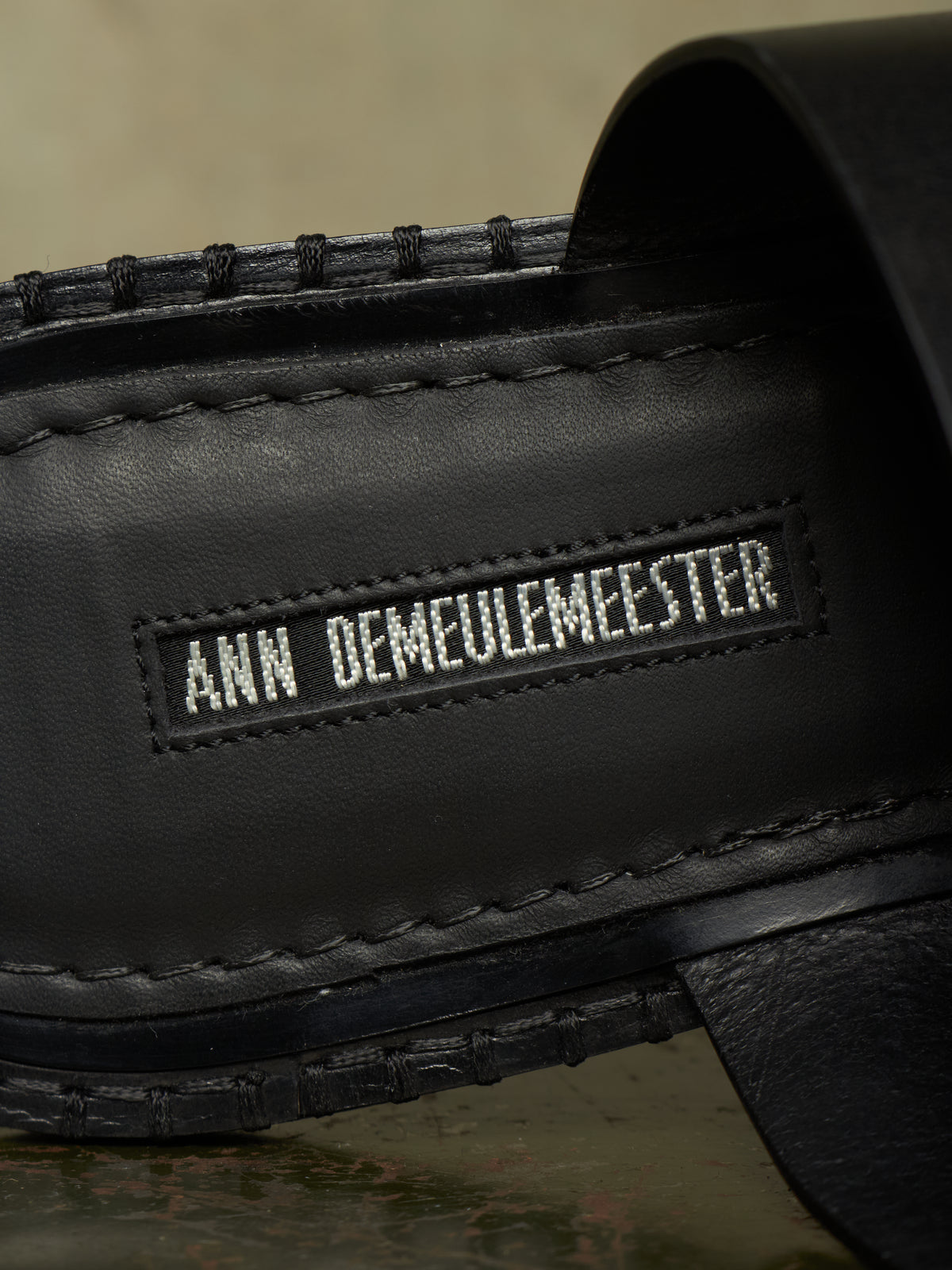 Close-up of a black leather shoe or sandal with visible stitching and a stitched brand label reading ANN DEMAULEMEESTER.