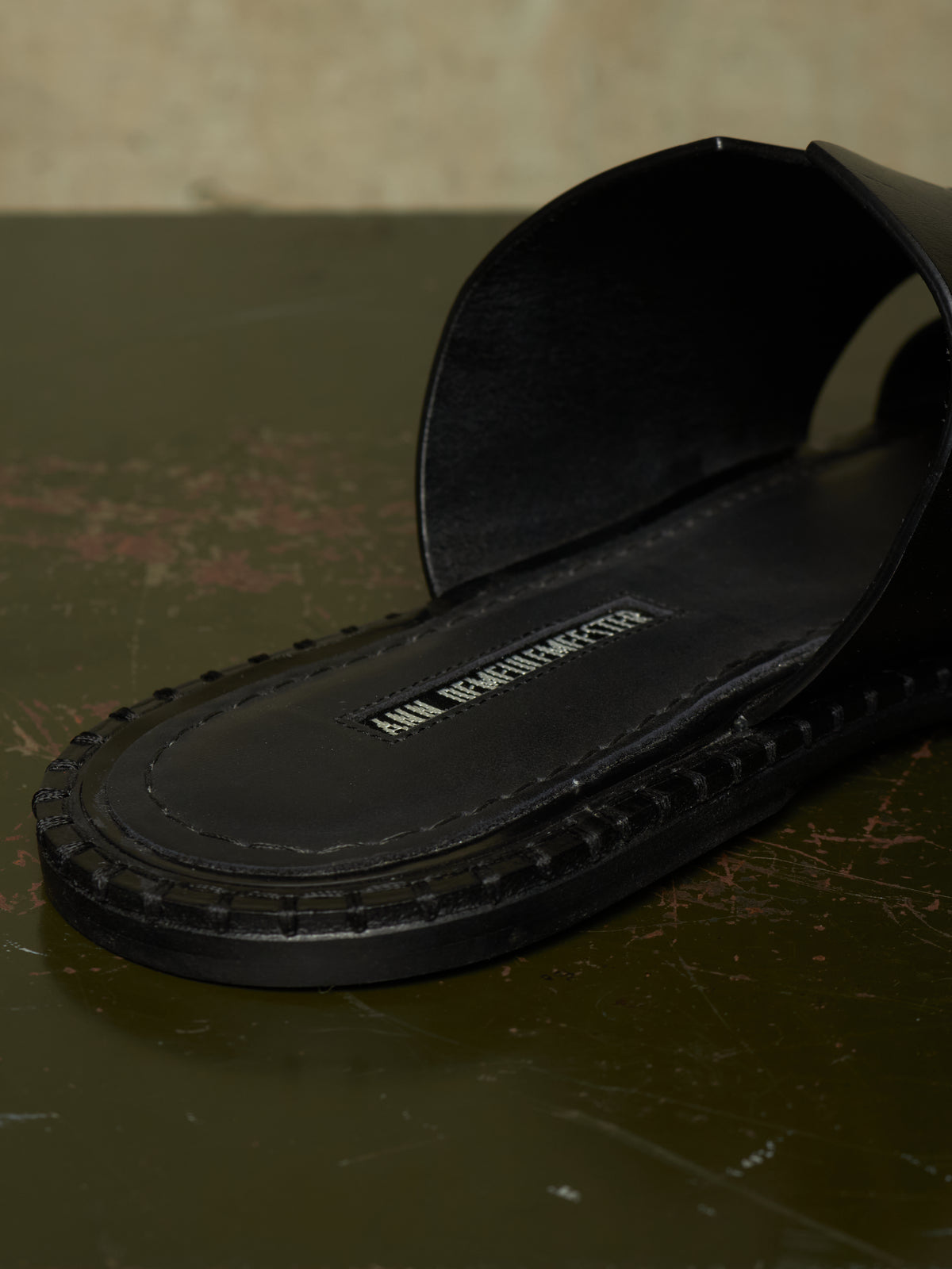 A close-up of a black leather slide sandal with a thick, textured sole, resting on a dark, rustic surface. The brand name 'ANN BERNHARDTS' is visible on the insole.