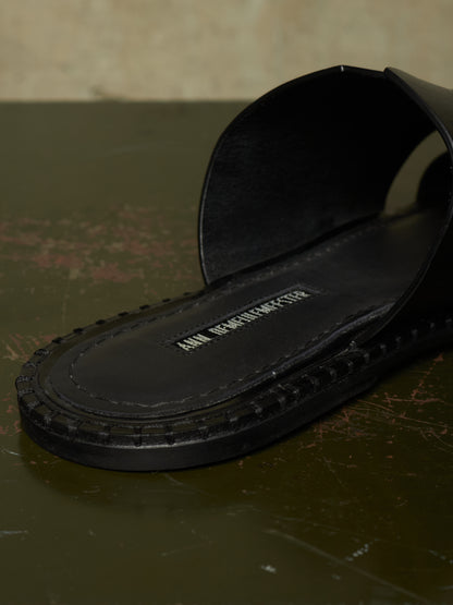 A close-up of a black leather slide sandal with a thick, textured sole, resting on a dark, rustic surface. The brand name 'ANN BERNHARDTS' is visible on the insole.