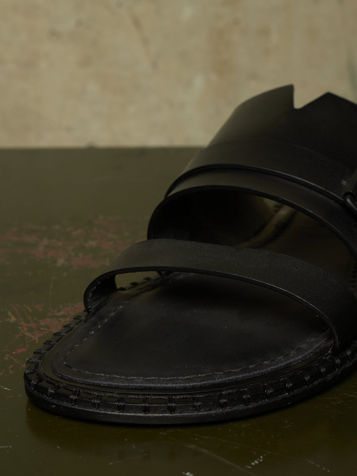 A close-up of a black leather slide sandal with wide straps and a textured sole, resting on a dark surface against a beige background.