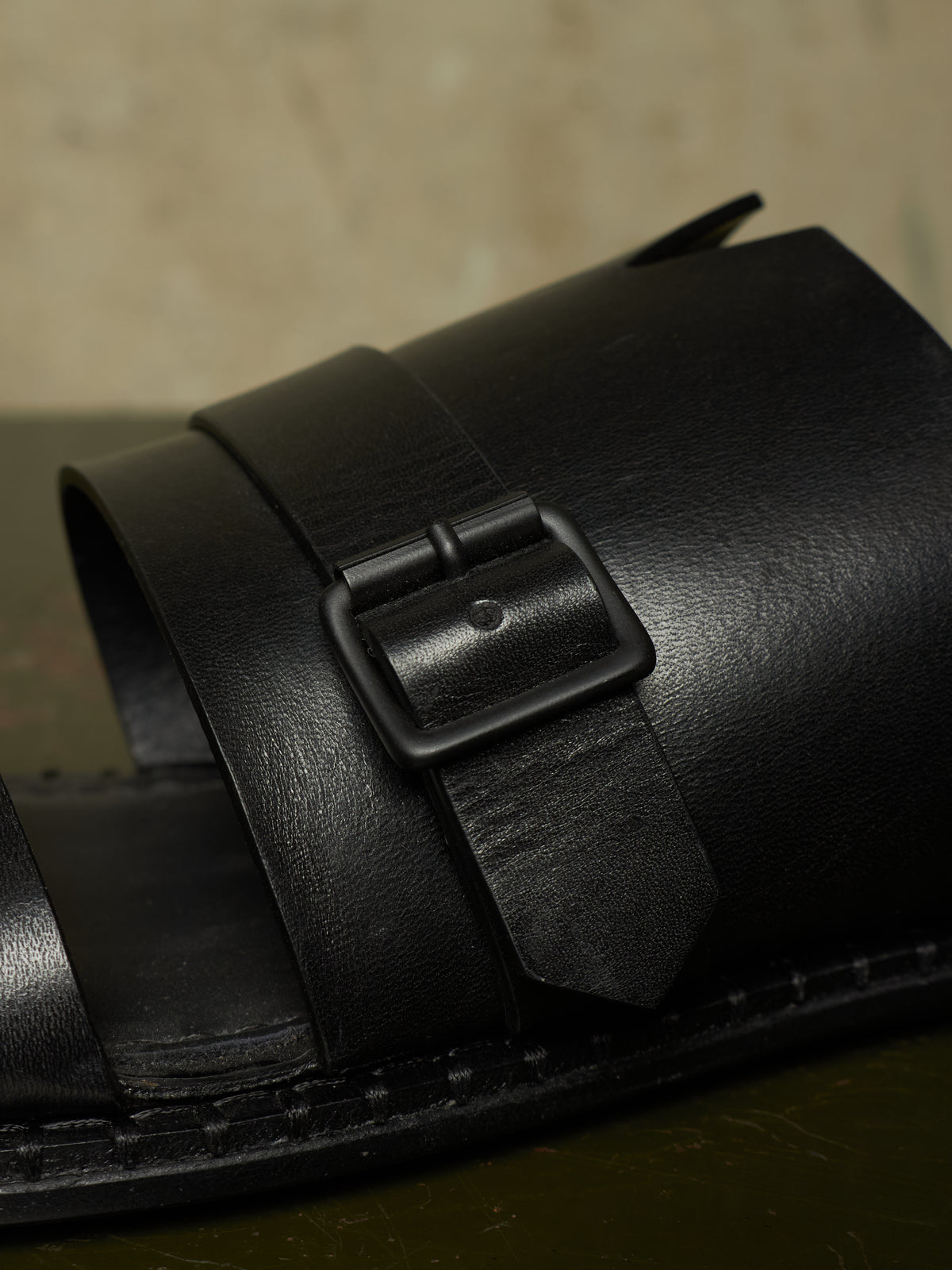 A close-up of a black leather double-strap sandal with a matte black buckle, resting on a dark surface against a textured beige background.