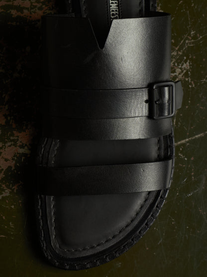A close-up of a black leather sandal with wide straps and a buckle, photographed on a dark textured surface.