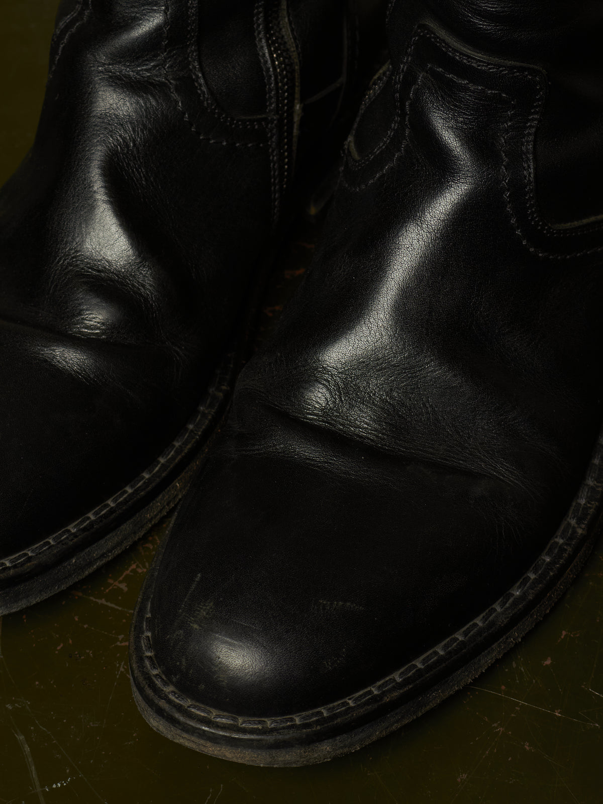 A close-up shot of a pair of black leather boots, showing the textured leather, stitching, and soles against a dark olive green background.
