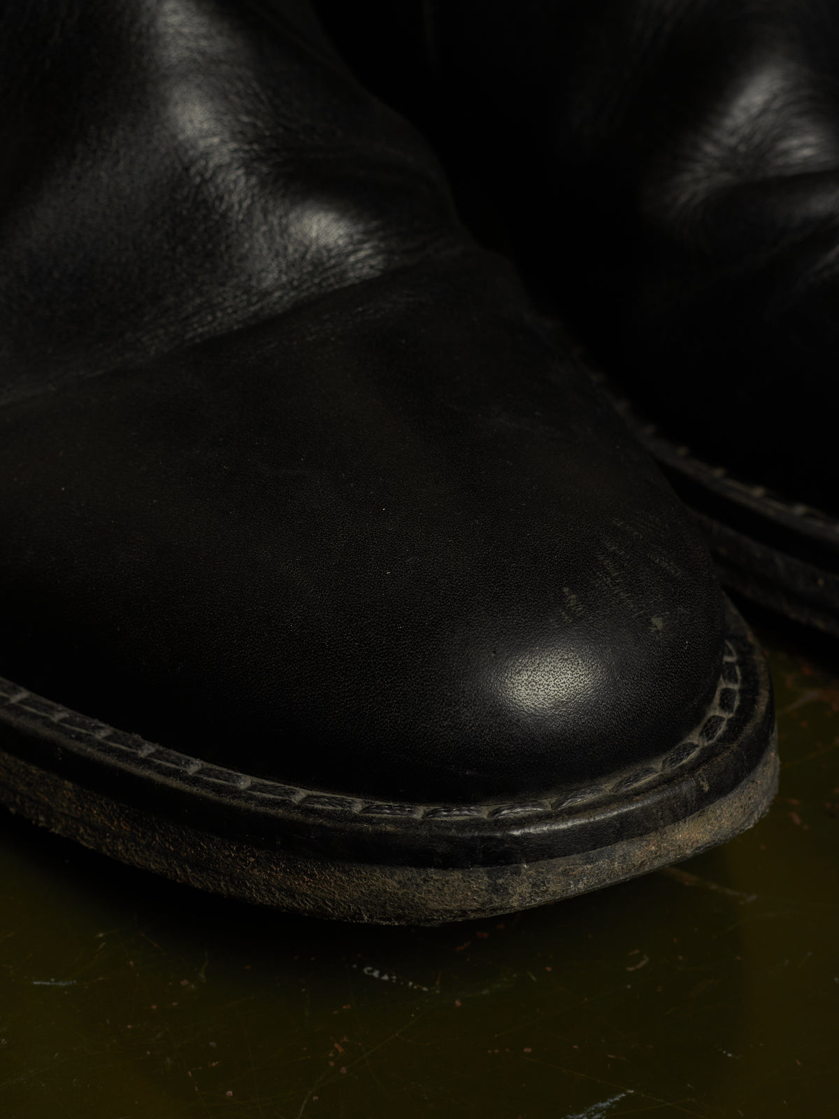 A close-up shot of a black leather boot toe showing the textured leather surface, decorative stitching around the sole, and a dark sole edge, with another boot partially visible in the background.