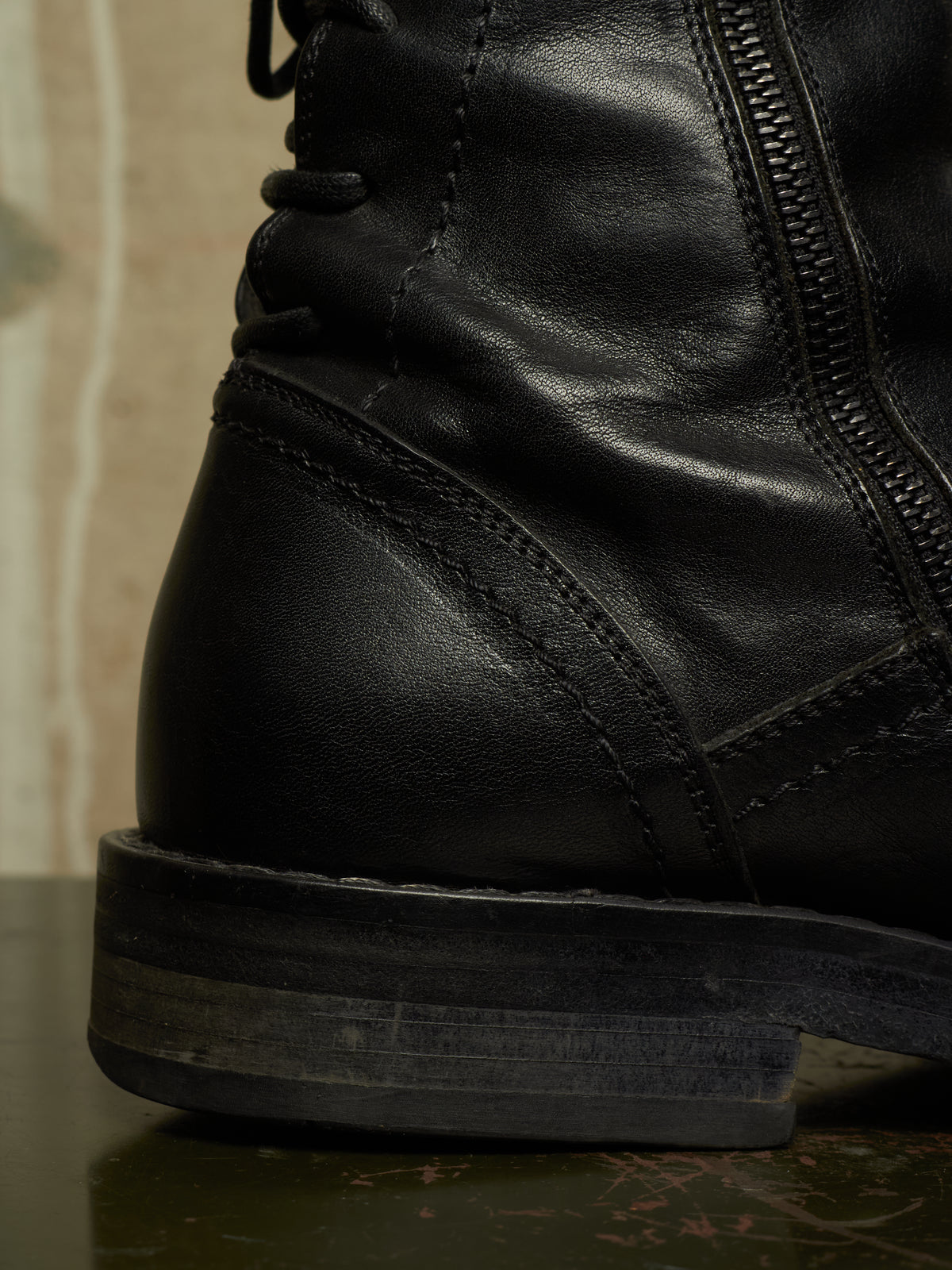 Close-up of a black leather lace-up boot showing the side zipper, stitching, and sole against a dark reflective surface.