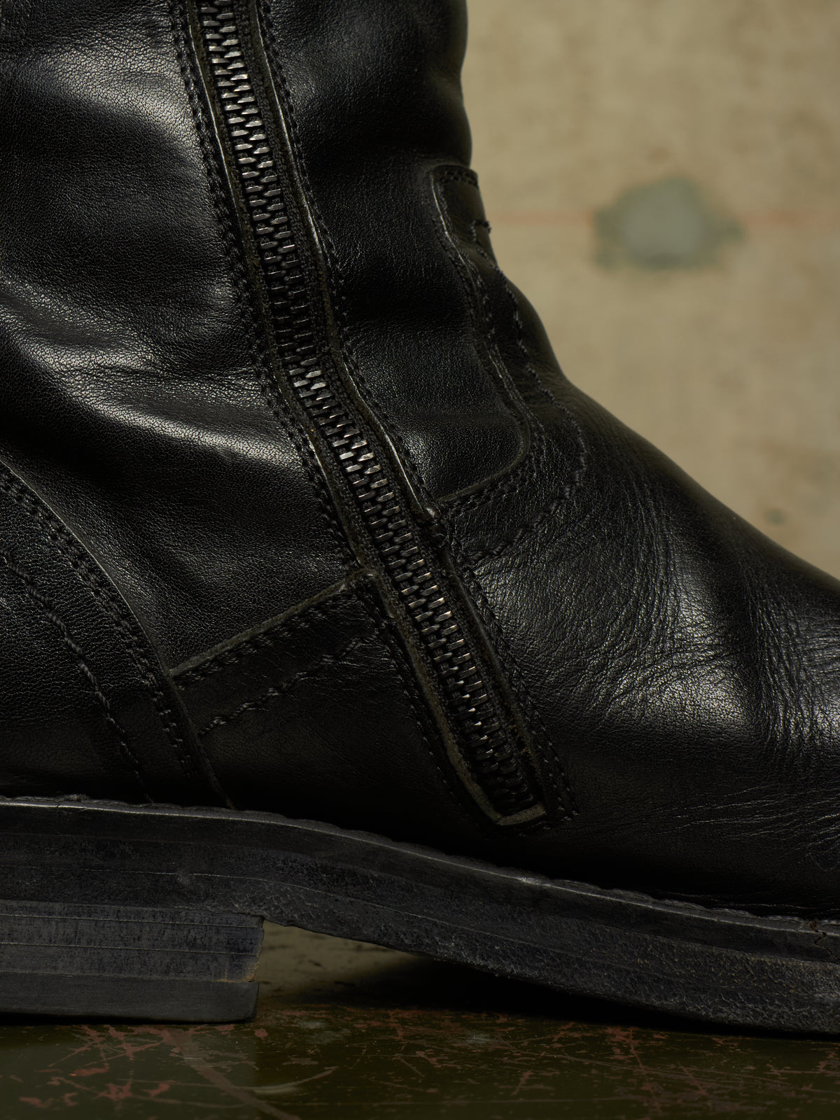 Close-up of a black leather boot with a side zipper, showing the texture of the leather and the zipper detail against a neutral background.