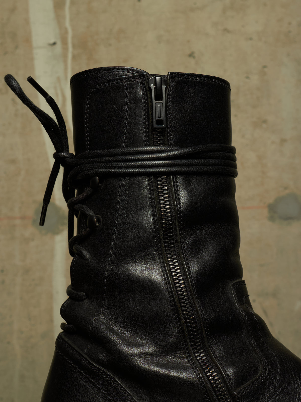Close-up of a black leather ankle boot with a side zipper, lace-up front, and a wrapped elastic band around the ankle, set against a textured concrete wall.