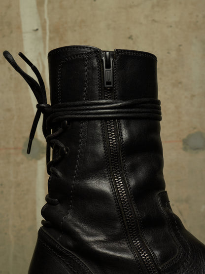 Close-up of a black leather ankle boot with a side zipper, lace-up front, and a wrapped elastic band around the ankle, set against a textured concrete wall.