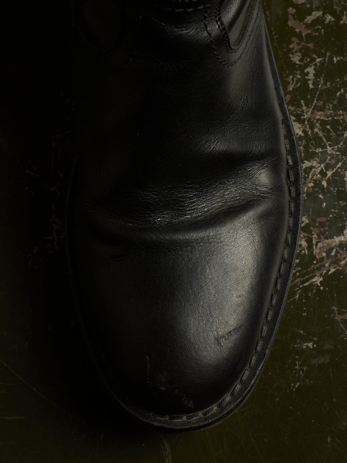 A close-up of a black leather boot against a dark, textured background.
