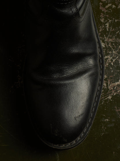 A close-up of a black leather boot against a dark, textured background.