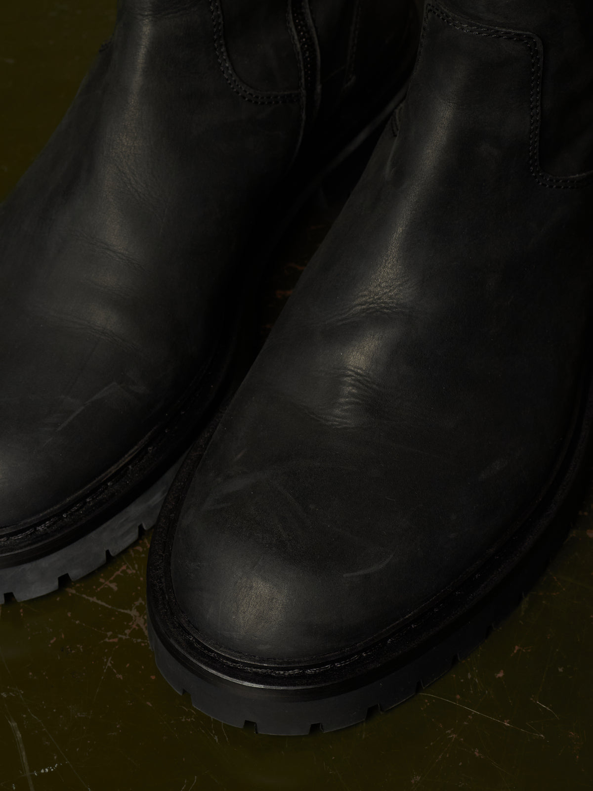 Close-up of a pair of black leather Chelsea boots with thick lug soles, resting on a dark olive green textured surface.