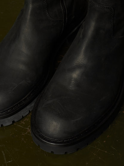 Close-up of a pair of black leather Chelsea boots with thick lug soles, resting on a dark olive green textured surface.