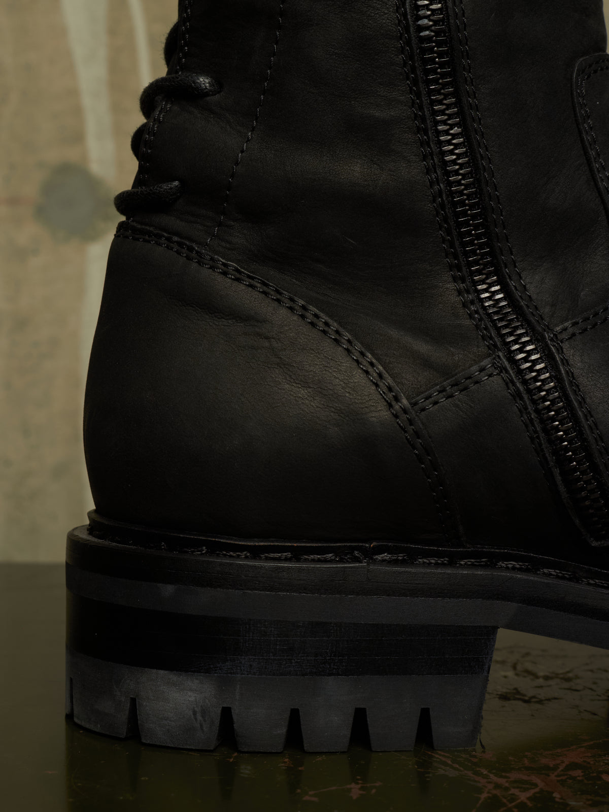 Close-up of a black leather lace-up boot with a side zipper and chunky lug sole, photographed against a dark textured background.