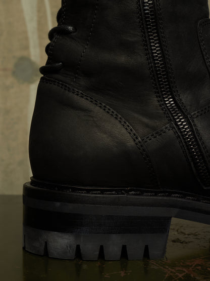Close-up of a black leather lace-up boot with a side zipper and chunky lug sole, photographed against a dark textured background.