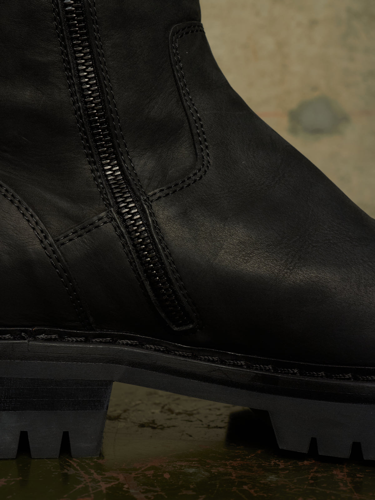 Close-up of a black leather boot with a side zipper, showing the texture of the leather and the rugged sole against a dark, moody background.