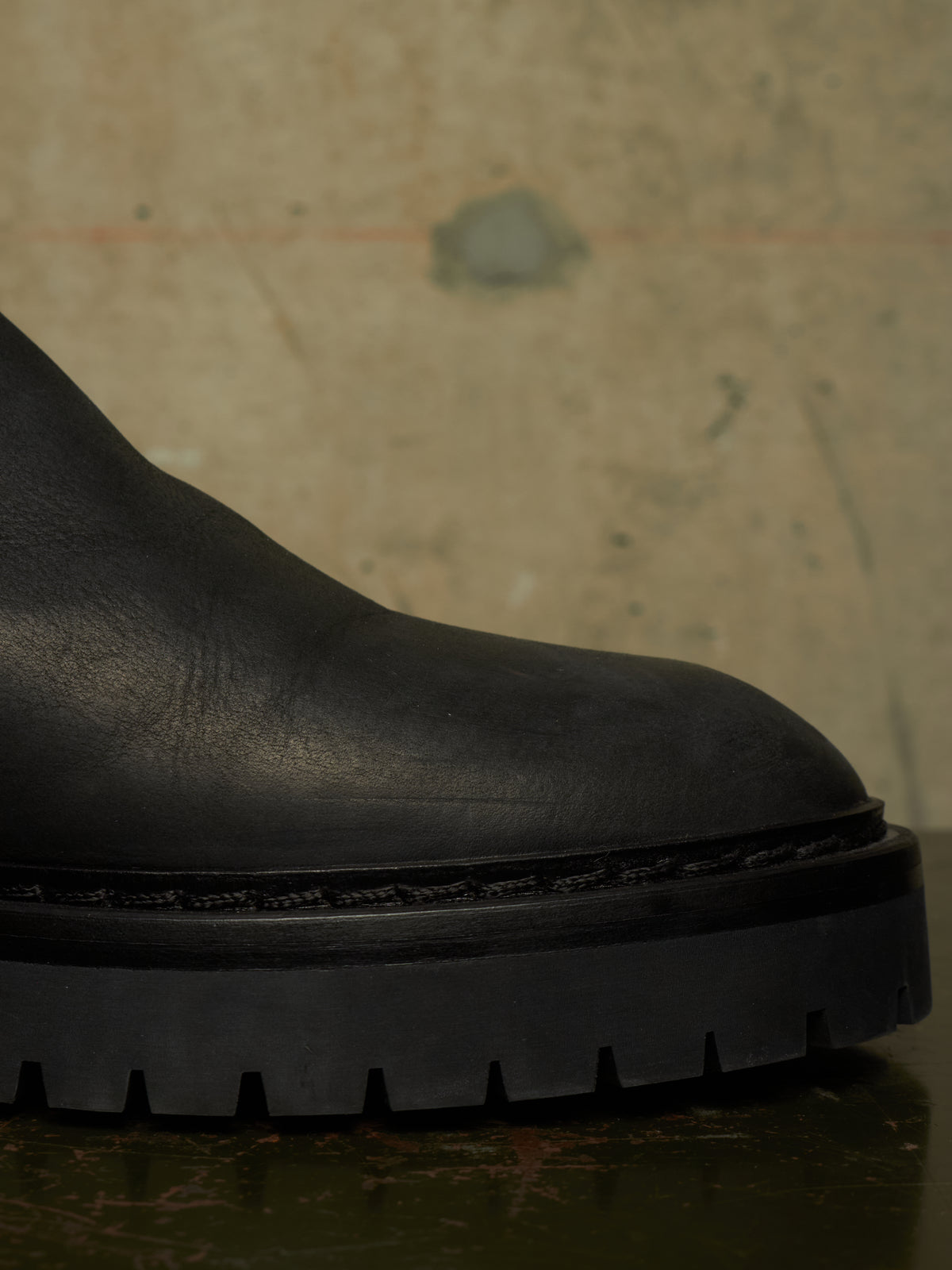 Close-up of a black leather boot with a chunky lug sole, resting on a dark reflective surface against a textured beige background.