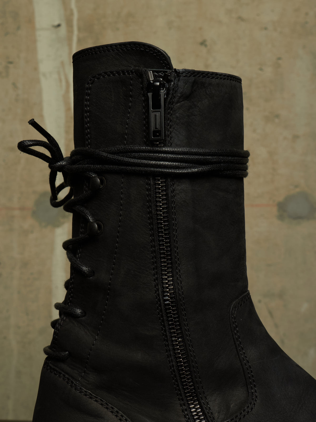 A close-up of a black leather lace-up ankle boot with a side zipper and a wrap-around strap detail near the top, set against a textured beige wall.