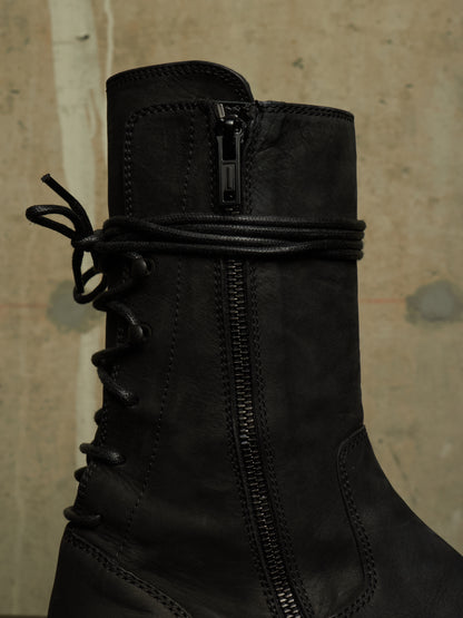 A close-up of a black leather lace-up ankle boot with a side zipper and a wrap-around strap detail near the top, set against a textured beige wall.