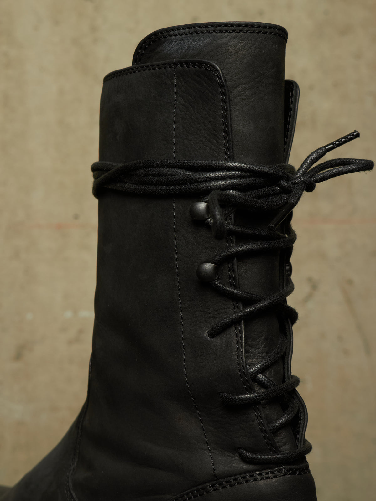 A close-up of a black leather lace-up boot against a textured beige background.