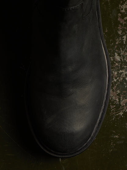 A close-up, top-down view of a black leather boot on a dark, textured green-brown surface.