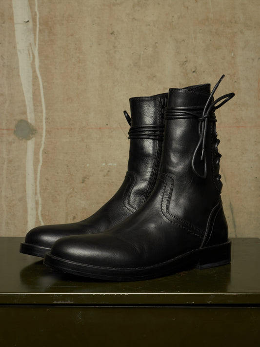A pair of black leather ankle boots with side zippers and lace-up detailing, displayed on a dark green surface against a textured beige wall.