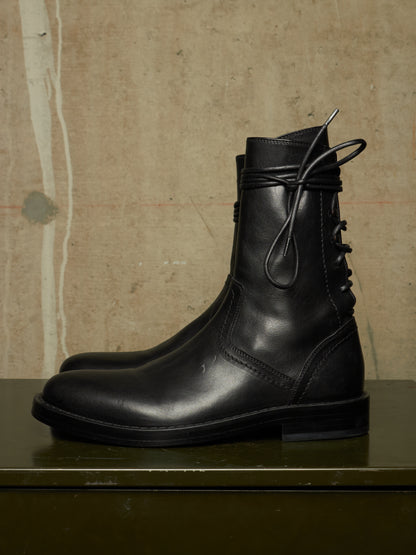 A pair of black leather mid-calf boots with lace-up detailing on the side, displayed on a dark green surface against a textured beige wall.