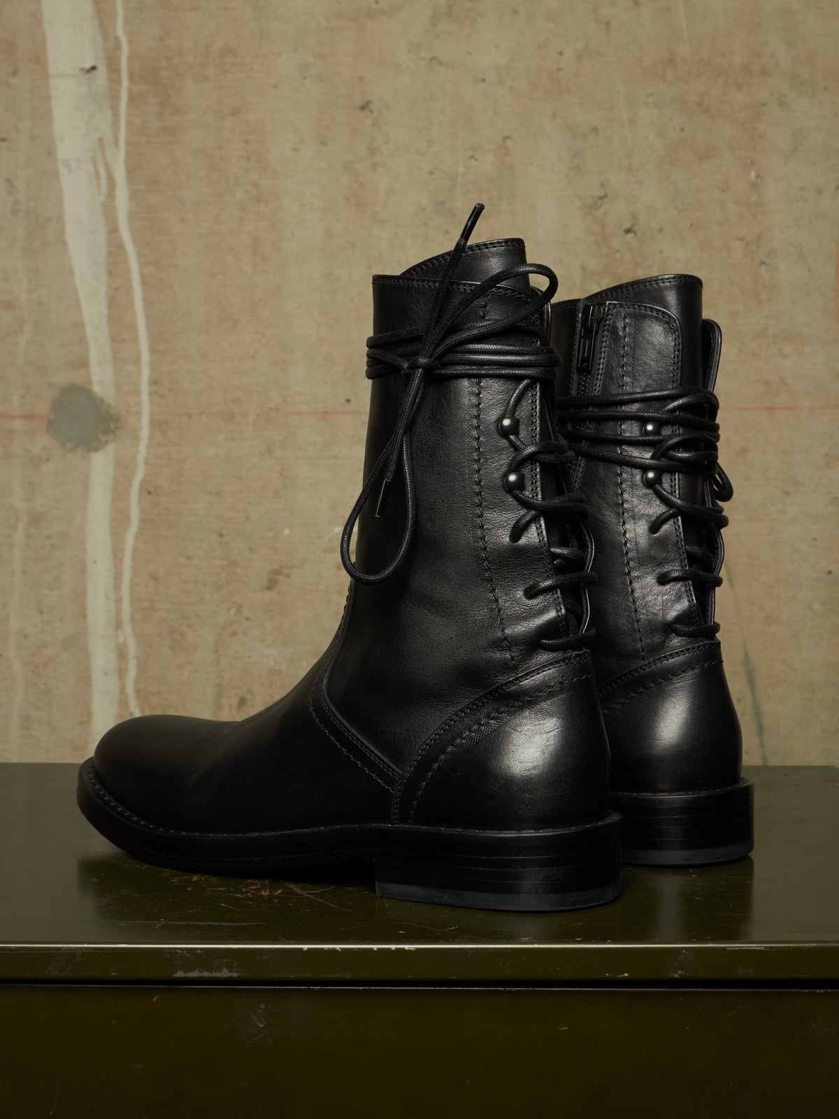 A pair of black leather lace-up ankle boots with a wraparound strap detail, displayed on a dark green surface against a textured beige wall.