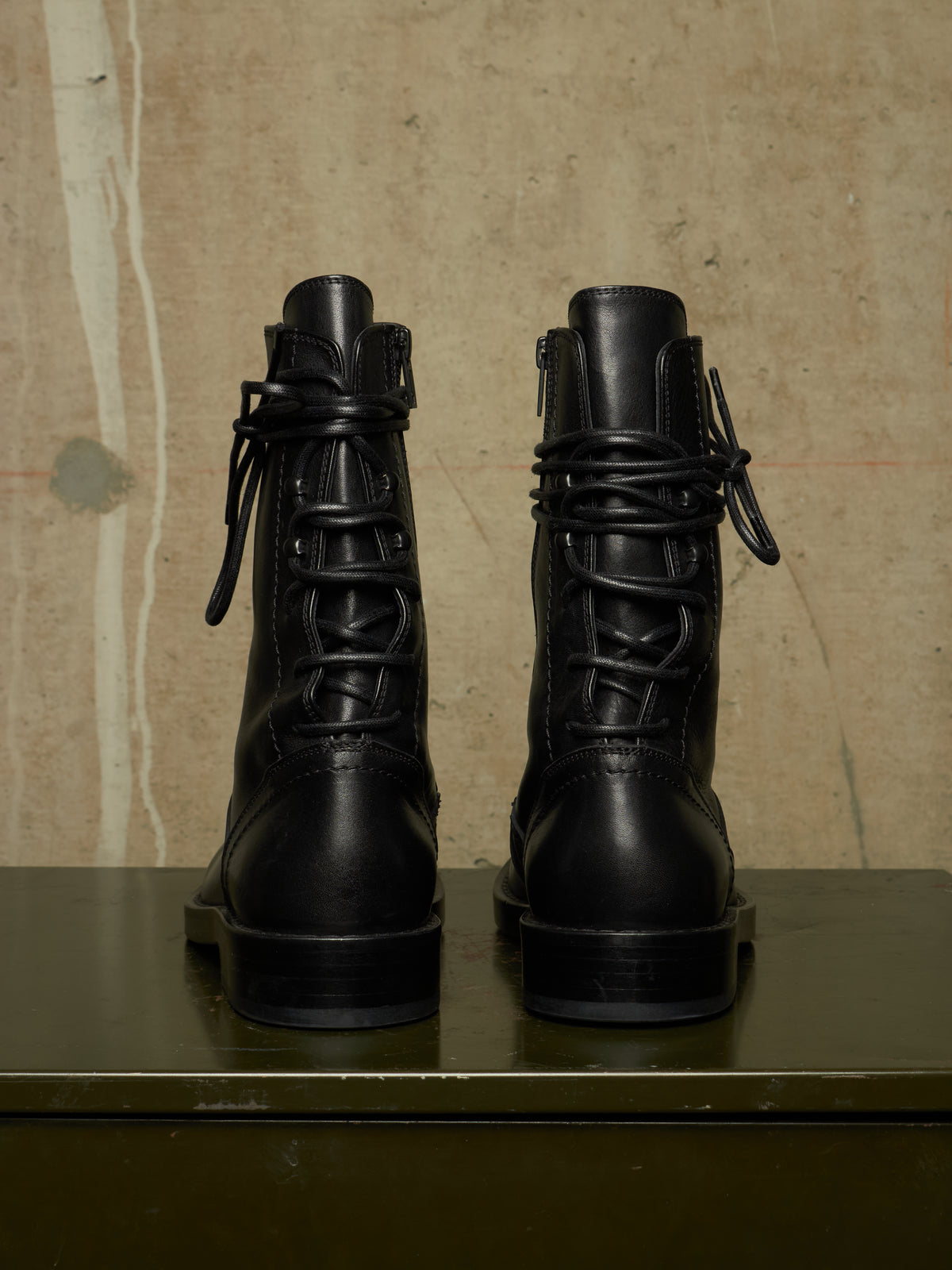 A pair of black leather lace-up combat boots shown from the back, resting on a dark green surface against a textured beige wall.