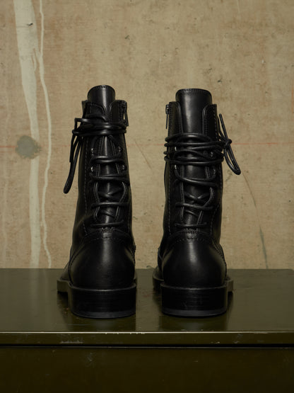 A pair of black leather lace-up combat boots shown from the back, resting on a dark green surface against a textured beige wall.