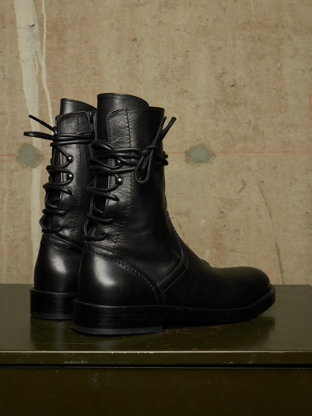 A pair of black leather lace-up ankle boots with a rugged sole, displayed on a dark green surface against a textured beige wall.