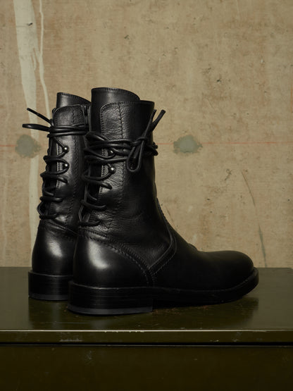 A pair of black leather lace-up ankle boots with a rugged sole, displayed on a dark green surface against a textured beige wall.