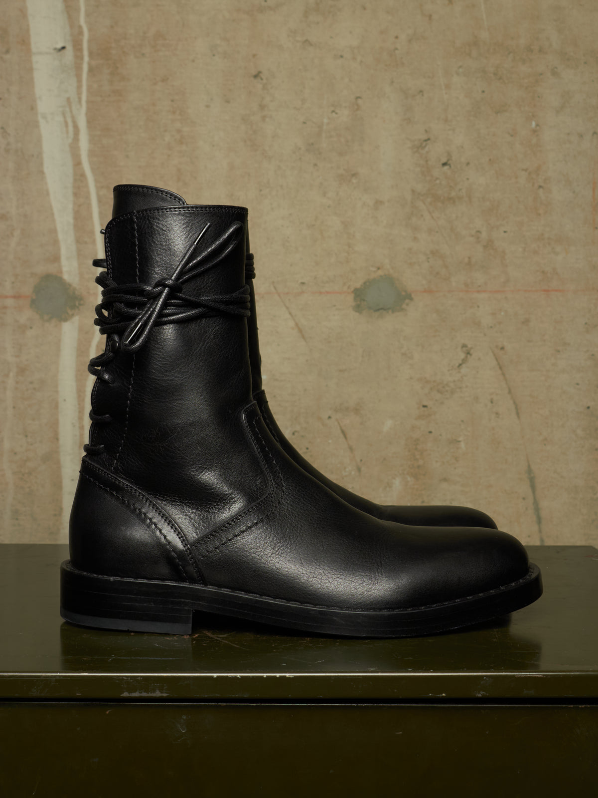 A black leather ankle boot with a decorative bow detail on the side, displayed on a dark green surface against a textured beige wall.