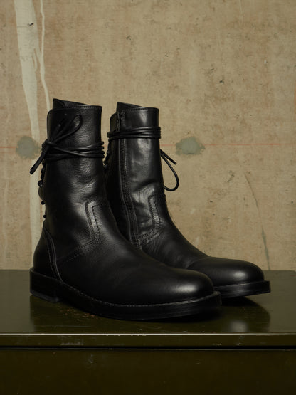 A pair of black leather ankle boots with lace-up front and side zippers, displayed on a dark green surface against a textured beige wall.