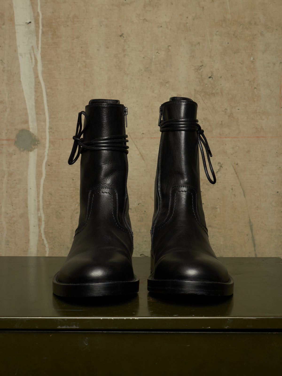 A pair of black leather ankle boots with side laces, displayed on a dark green shelf against a textured beige concrete wall.