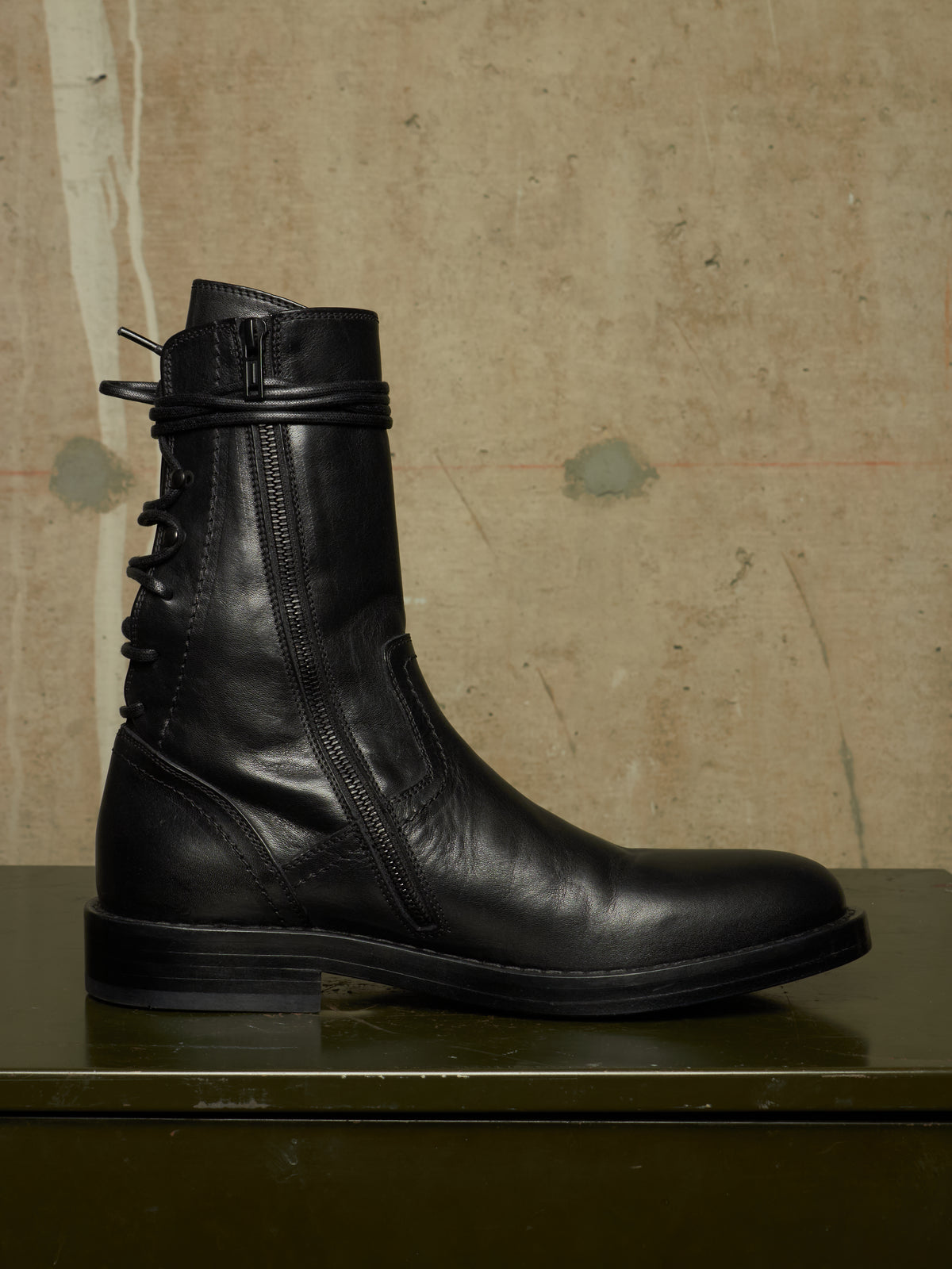 A black leather ankle boot with a side zipper, lace-up front, and decorative strap detail near the collar, displayed on a dark green surface against a textured beige wall.