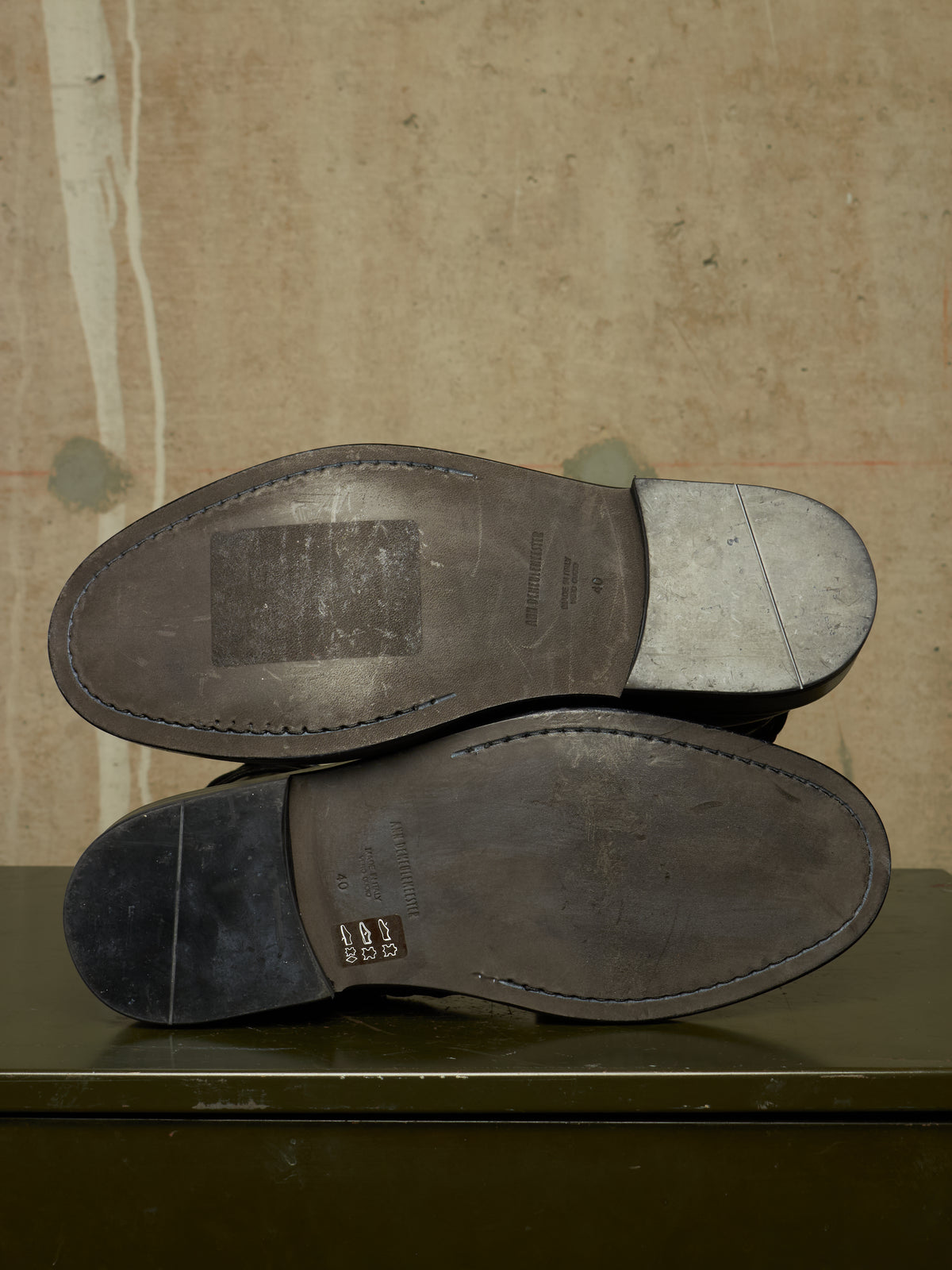 The soles of a pair of dark leather shoes are shown from underneath, resting on a dark green surface against a textured beige background.