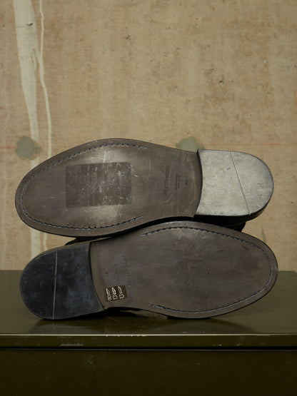The soles of a pair of dark leather shoes are shown from underneath, resting on a dark green surface against a textured beige background.