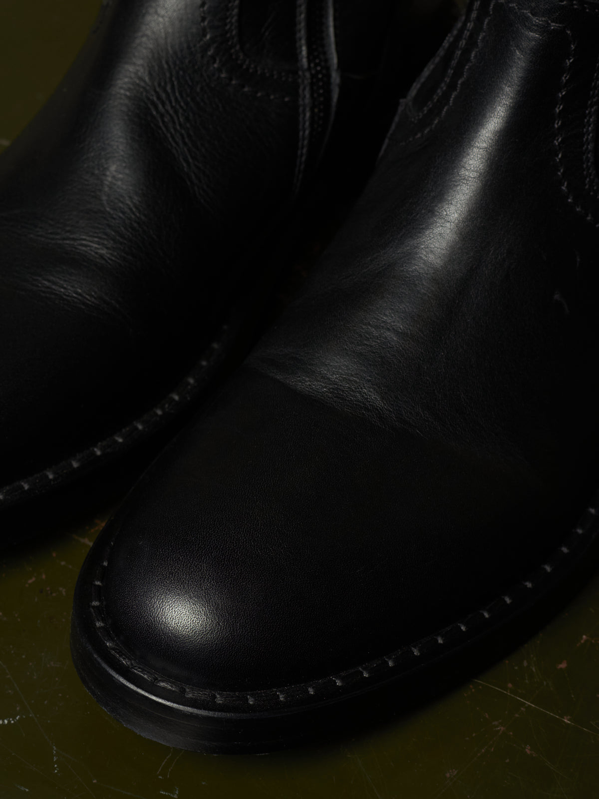 Close-up of a black leather ankle boot showing the toe cap, stitching, and side elastic gore on a dark olive background.