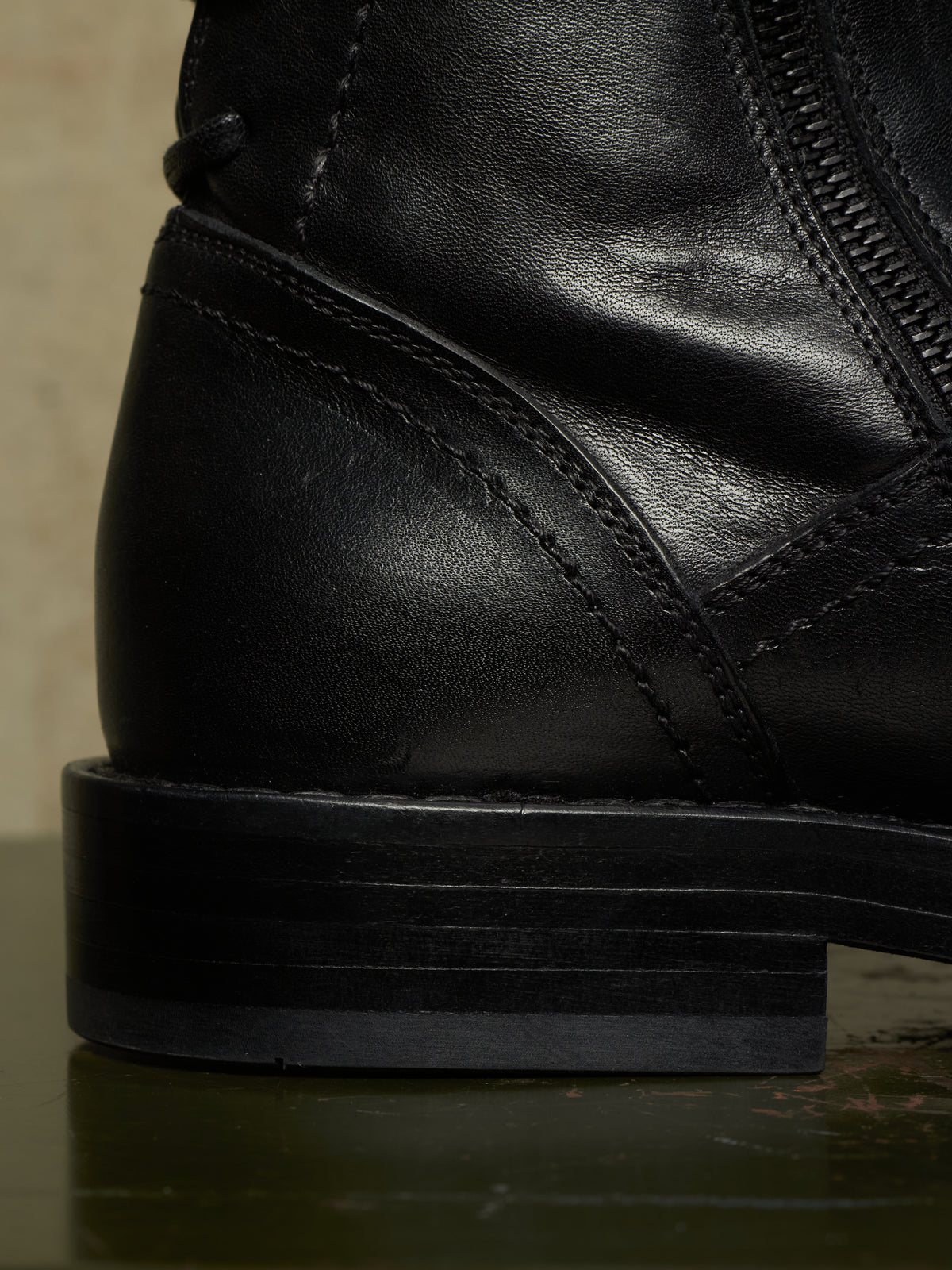 Close-up detail shot of a black leather ankle boot showing the heel, side stitching, zipper, and stacked black sole.