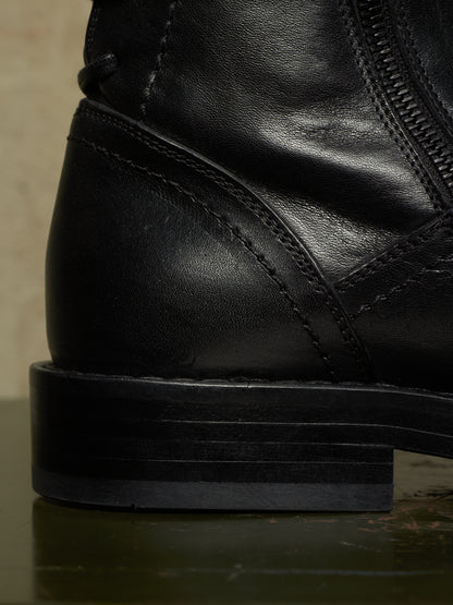 Close-up detail shot of a black leather ankle boot showing the heel, side stitching, zipper, and stacked black sole.