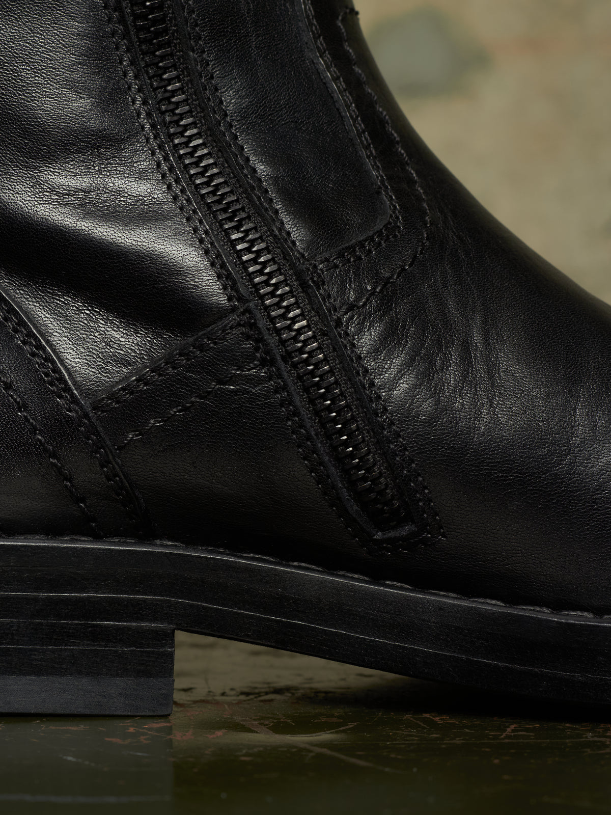 Close-up of a black leather ankle boot with a side zipper and stacked sole, photographed against a dark textured background.