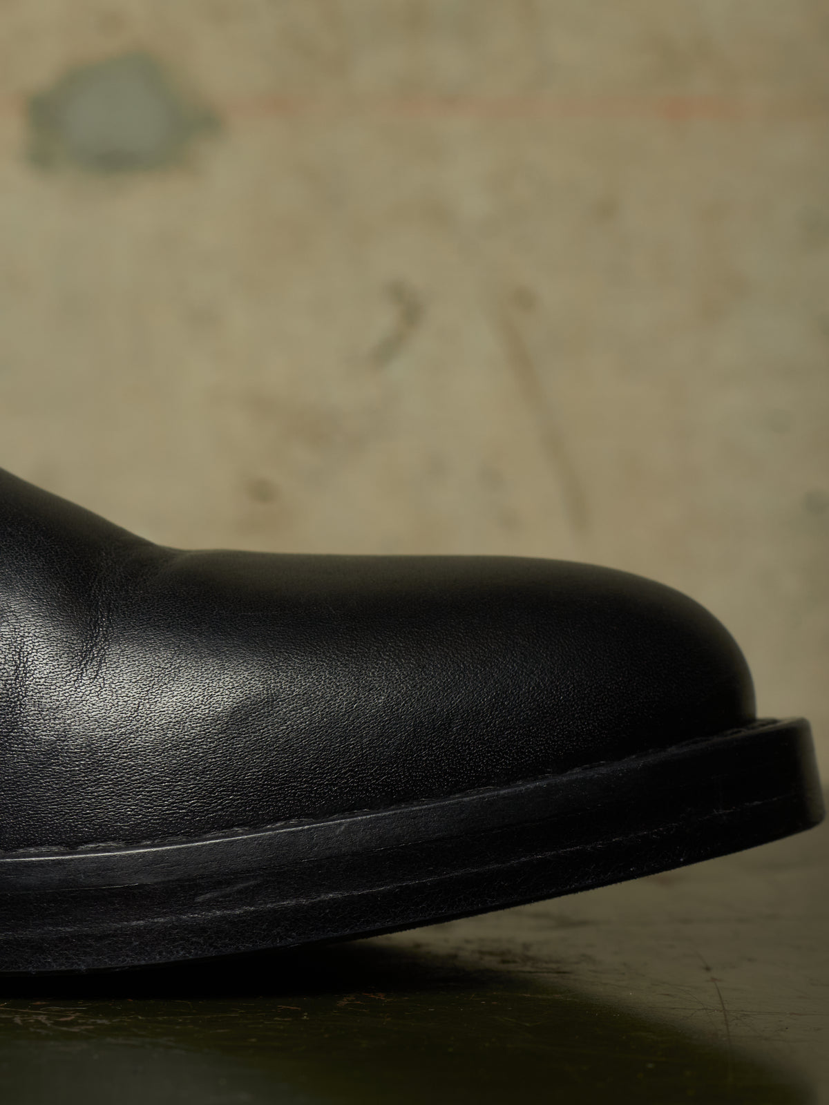 Close-up of the rounded toe and side of a black leather boot against a textured beige background.