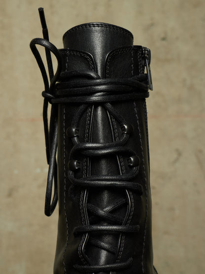 Close-up of a black leather lace-up boot with a unique twisted cord detail wrapping around the eyelets, set against a textured beige background.