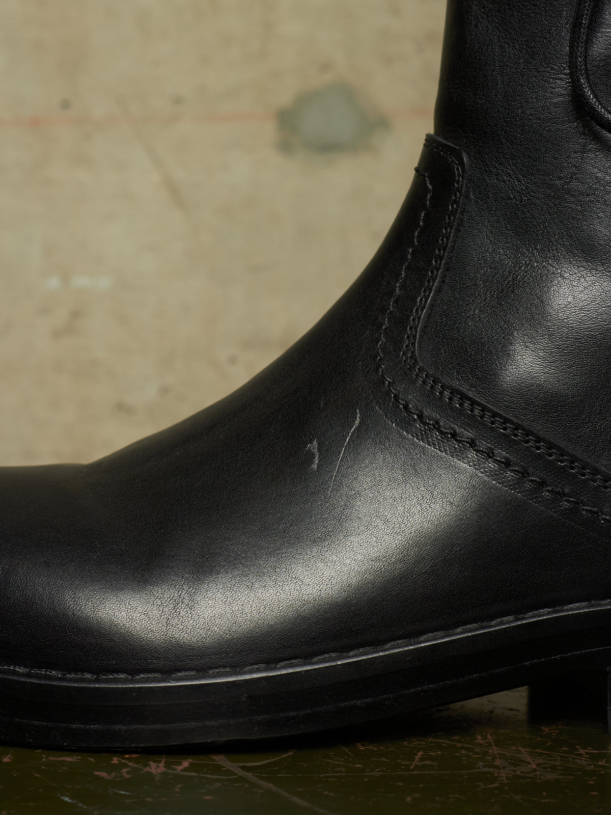 A close-up shot of a black leather boot, showing the side stitching and toe area against a textured, warm-toned background.