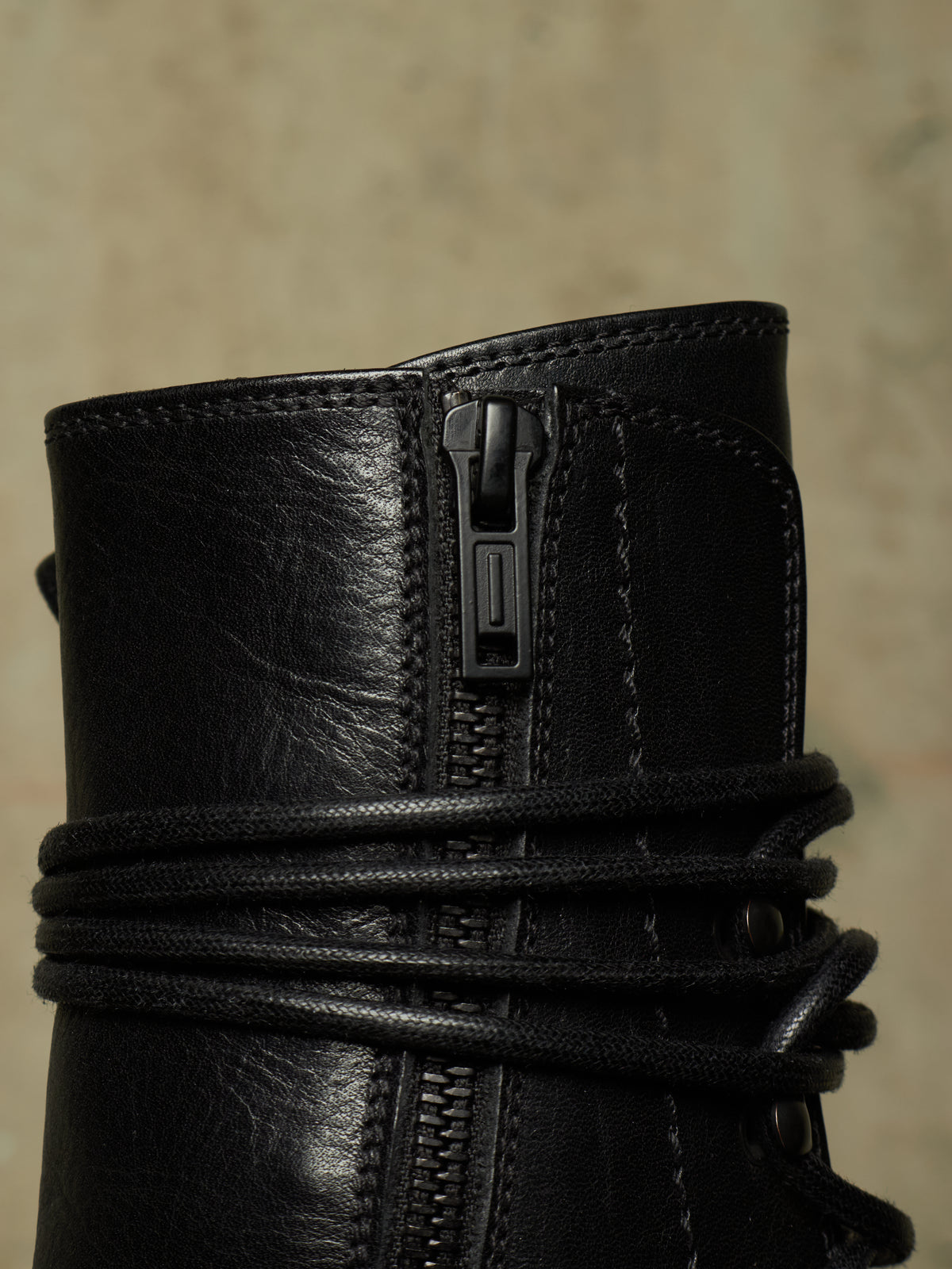 Close-up of a black leather boot featuring a central zipper and wrapped black laces, set against a textured beige background.