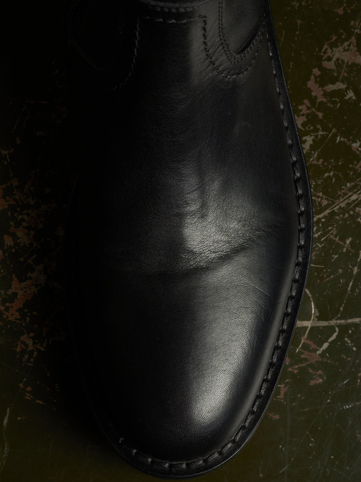 A close-up of a black leather boot toe showing the smooth leather texture, whipstitching along the side, and a dark textured background.