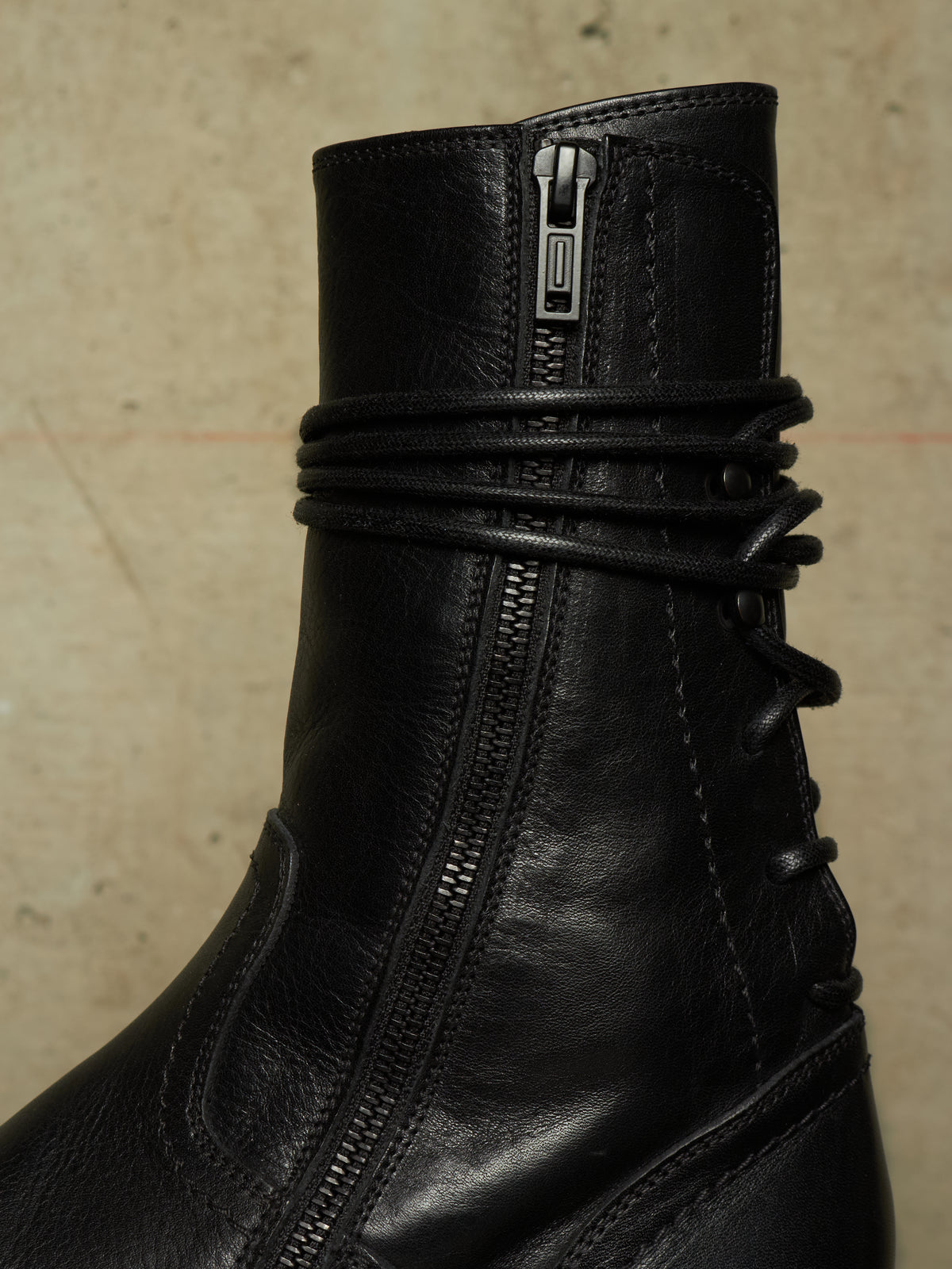 Close-up of a black leather ankle boot featuring a side zipper, a wrap-around lace detail, and a pull tab at the top, set against a neutral textured beige background.
