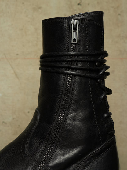 Close-up of a black leather ankle boot featuring a side zipper, a wrap-around lace detail, and a pull tab at the top, set against a neutral textured beige background.