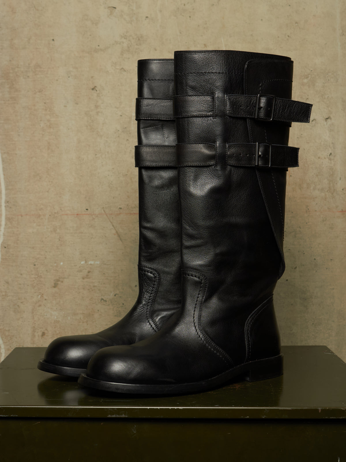 A pair of tall black leather boots with multiple decorative strap details around the shaft, displayed on a dark surface against a textured beige wall.