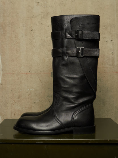 A pair of black leather knee-high riding boots with double buckle straps on the shaft, displayed on a dark green surface against a textured beige wall.